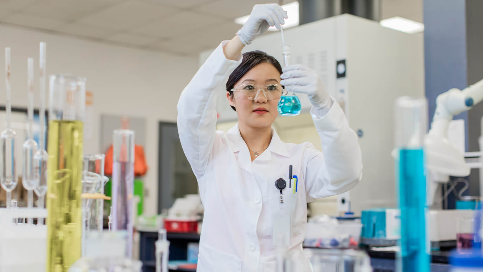 Curious scientist wearing goggles and using dropper to mix beaker sample in laboratory experiment.